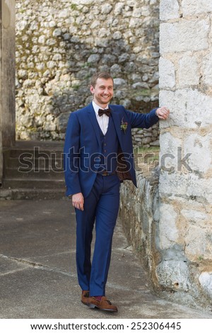handsome groom in a blue suit smiling leaning on the wall