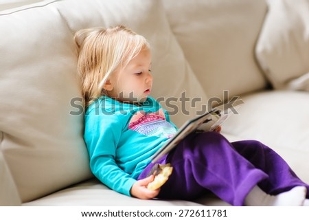 Cute little baby or toddler girl reading a book on a leather couch indoors on a rainy day