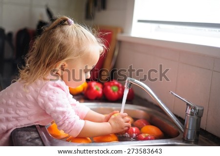Cute little child, a toddler girl with blonde curly hair helping by washing healthy fruit and vegetables in a sink indoors in a kitchen