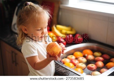 Cute little child, a toddler girl with blonde curly hair helping by washing healthy fruit and vegetables in a sink indoors in a kitchen