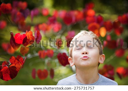 An outdoor portrait of a cute little boy blowing or sending a kiss into a camera with his eyes shut with a pretty tree with red heart shaped leaves in the background