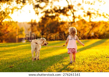 Cute little child, adorable girl with curly hair and her dog, yellow labrador walking away into the sunset light in a countryside park