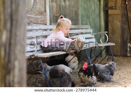 Adorable little toddler girl feeding birds chickens in a barn sitting on a wooden bench