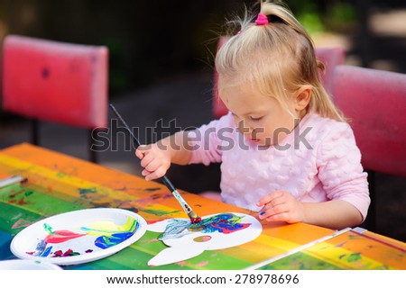 Cute little toddler girl drawing on a mask with paint on a colourful table