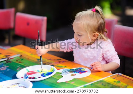Cute little toddler girl drawing on a mask with paint on a colourful table