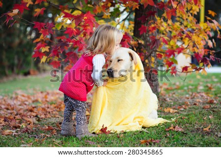Adorable blonde little girl kissing her dog a yellow labrador wrapped in a cozy warm blanket in a park with autumn leaves in the background Adorable blonde little girl kissing her dog a yellow labrador wrapped in a cozy warm blanket in a park with autumn leaves in the background