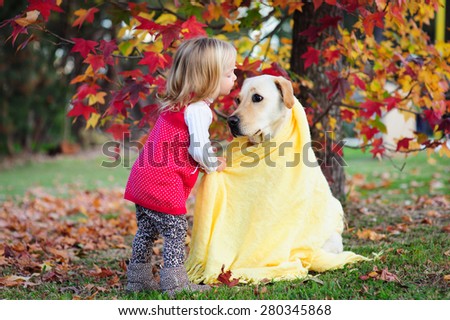 Adorable blonde little girl kissing her dog, a yellow labrador wrapped in a cozy warm blanket in a park with autumn leaves in the background