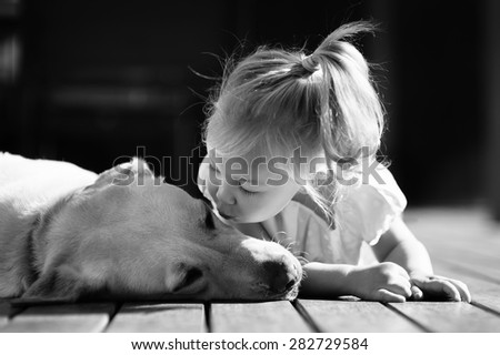 A black and white portrait of a little cute toddler girl kissing her dog, a yellow labrador relaxing on the wooden deck in the sun