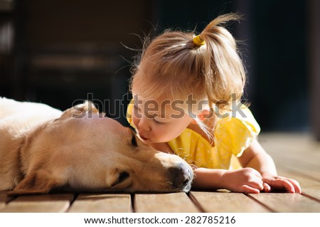 A portrait of a little cute toddler girl kissing her dog, a yellow labrador relaxing on the wooden deck in the sun