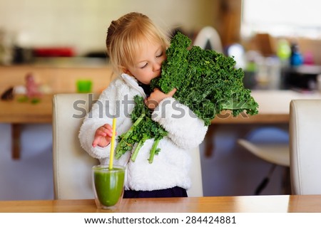 Little toddler girl in a white fluffy jacket holds a bunch of fresh kale and is about to drink a green smoothie in the kitchen