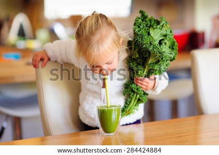 Little toddler girl in a white fluffy jacket holds a bunch of fresh kale and is about to drink a green smoothie in the kitchen