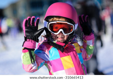 Cute little girl wearing a bright colorful winter jacket and a pink ski helmet and goggles waiving at the camera