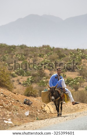 SIDI IFNI, MOROCCO, AUGUST 29: Moroccan man sitting on his donkey and walking along the road towards Guelmim with mountains and arid vegetation in the background. Morocco 2014.