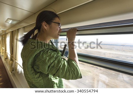 LHASA, CHINA, APRIL 15: Chinese young woman looking by an open window and holding her camera in the Train Shanghai-Lhasa. Across the window, we can see  some Tibetan landscape. China 2013.