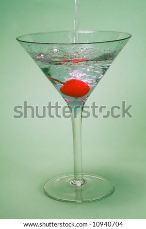 cocktail glass with drink being poured in with a cherry in the glass on a green background