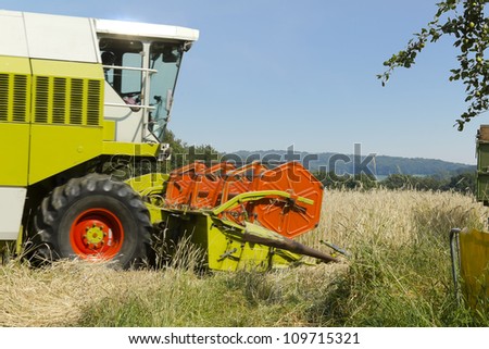 Harvester in a field of wheat bringing in the crop. Subtle motion blur on wheels and combine. Hills and blue sky in the background. An apple tree is seaming the upper right corner.