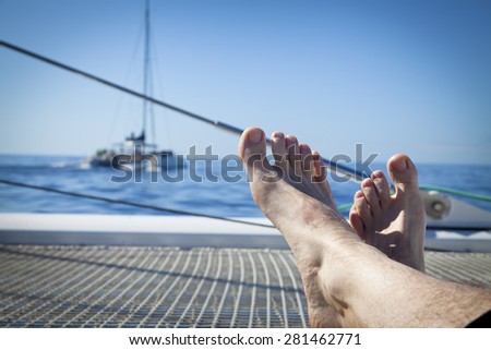 man lounging on a catamaran sailboat trampoline with her feet propped up and crossed. calm blue ocean and cloudless blue sky are in the background. copy space available