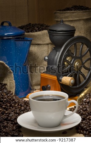 Coffee cup propped with coffee pot, coffee beans, burlap bags of coffee and a vintage coffee grinder