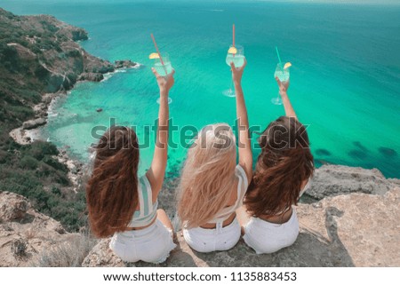 Cheers! Back view of Beautiful traveller girl friends having fun with coctail glass on cliff mountains enjoying lagoon bay over Fiolent, Crimea. Three young women resting on a beach. Travel lifestyle.