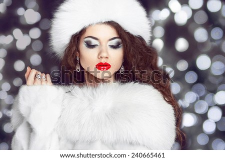 Fashion girl model posing in fur coat and white furry hat. Winter Beautiful Woman in Luxury clothes over bokeh Christmas Lights background.