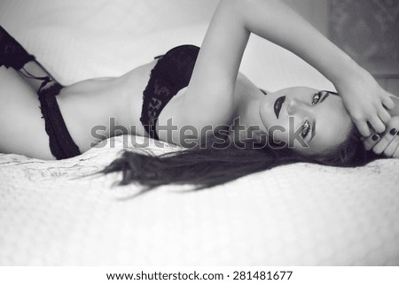Alluring slim girl in black lingerie lying on bed. Brunette woman with long wavy hair in sexy bra and panties. Black and white portrait.