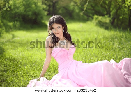 Happy smiling young bride girl dreaming and resting on green grass at spring park. Attractive brunette model wearing in pink wedding dress.