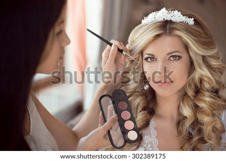 Professional Stylist makes makeup bride on the wedding day. Beautiful smiling blond woman with long curly hair style.