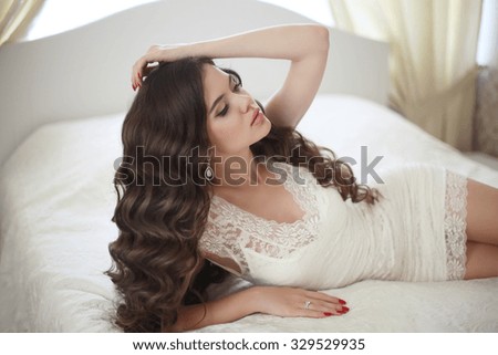 Hairstyle. Beautiful brunette bride girl with long healthy wavy hair styling and professional makeup in short lace wedding dress lying in white bed posing in modern interior.