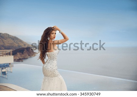 Beautiful bride girl in beaded wedding dress. Summer holiday fashion concept. Luxury resort woman posing by infinity swim pool over blue sky with sea.