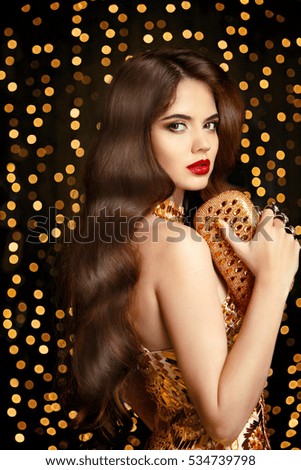 Elegant fashion brunette woman. Wavy hair style. Red lips Makeup. Healthy shiny hairstyle. Sexy girl model in golden dress with trendy clutch bag Accessories over bokeh lights background.