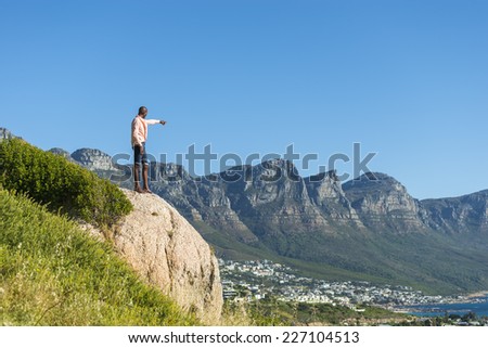African black man, standing on a high rock overlooking Cape Town as he points and scouts the blue sky, ocean and mountains on a sunny summers day