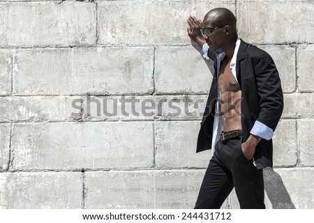 African black man model with six pack in black suit and unbuttoned white shirt, wearing sun glasses on beach with concrete wall background