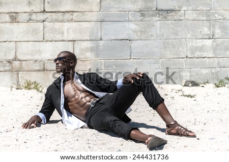 African black man model with six pack in black suit and unbuttoned white shirt, wearing sun glasses on beach with concrete wall background