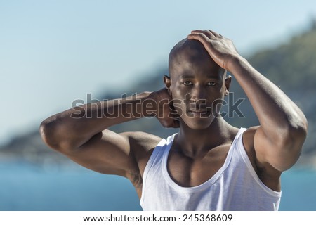Standing African black man wearing white vest and blue short jeans. Male model thinking while isolated alone by a blue ocean and sky background. Cape Town South Africa