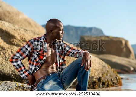 African black man model with six pack in unbuttoned checkered shirt, isolated against a beach rocks, sand and blue sky