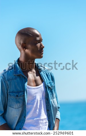 African black man wearing white vest and blue jean shirt with short jeans. Male model thinking while isolated alone by a blue ocean and sky background. Cape Town South Africa