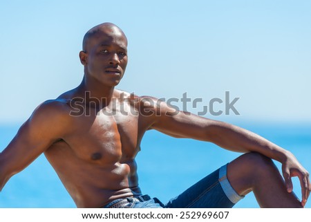Topless African black man sitting with short jeans. Male model thinking while isolated alone by a blue ocean and sky background. Cape Town South Africa