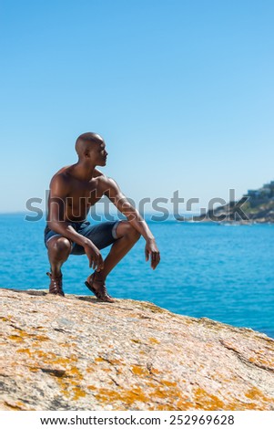 Topless African black man crouching with short jeans. Male model thinking while isolated alone by a blue ocean and sky background. Cape Town South Africa