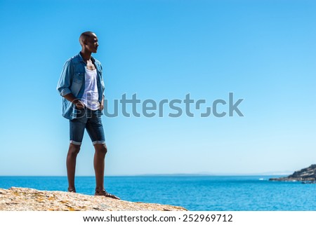 African black man wearing white vest and blue jean shirt with short jeans. Male model thinking while isolated alone by a blue ocean and sky background. Cape Town South Africa