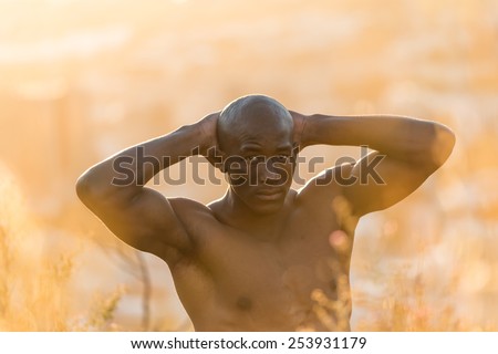 African american black male model standing topless with no shirt, showing muscular fit body at sunrise in long grass and bush