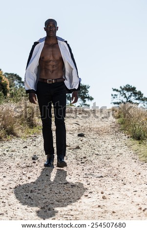 African black male model, wearing black suit with white unbuttoned shirt, walking down gravel path outdoors in nature with mountain views.