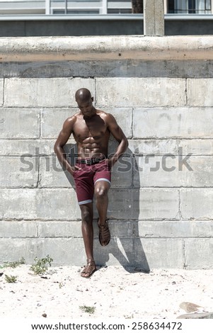 Topless, fit muscular african american male model isolated against a white concrete wall, wearing maroon shorts.