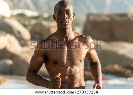 Topless, athletic, muscular and healthy black man running along the beach, splashing water during sunset
