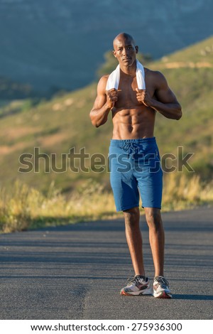 Athletic, sporty, muscular, healthy black male undressing by taking his white vest off, along a road outdoors with a mountain background while resting