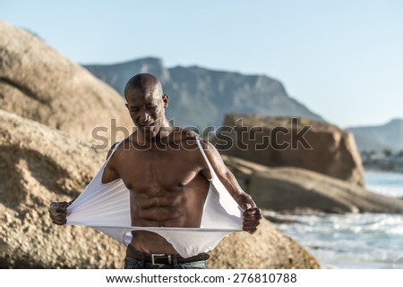 Muscular African american black male model, ripping his white vest shirt on the beach with great power and anger