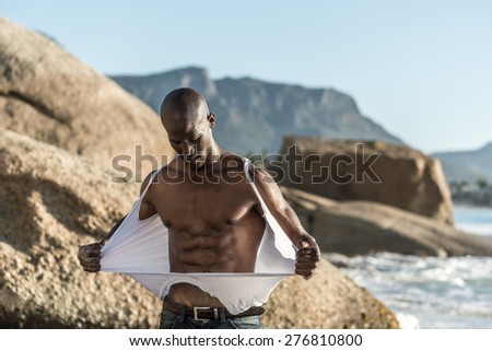 Muscular African american black male model, ripping his white vest shirt on the beach with great power and anger