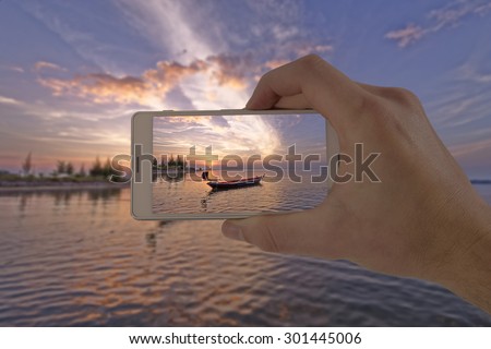 Tourist hand holding smart phone, taking photo of Thailand beach sunset boat