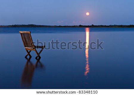Singular chair in calm water facing the land in the horizon. With rising orange moon reflected in the blue water