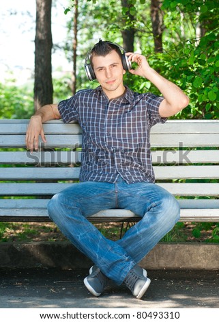 Portrait of a relaxed young man sitting on bench in park and listening to music on headphone