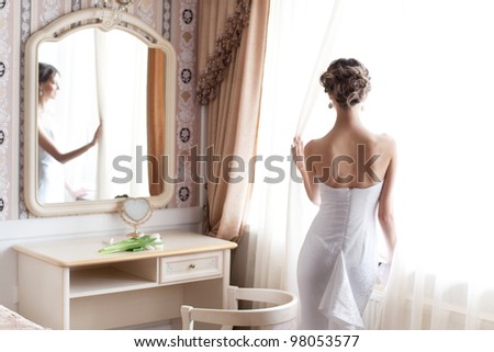 Beautiful bride in white wedding dress standing in her bedroom near the window